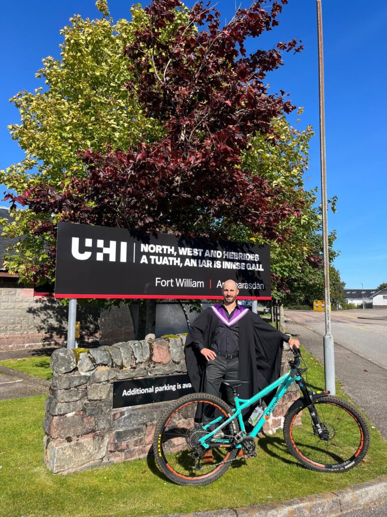 Graduate in cap and gown posing with a turquoise mountain bike in front of the UHI Fort William sign.