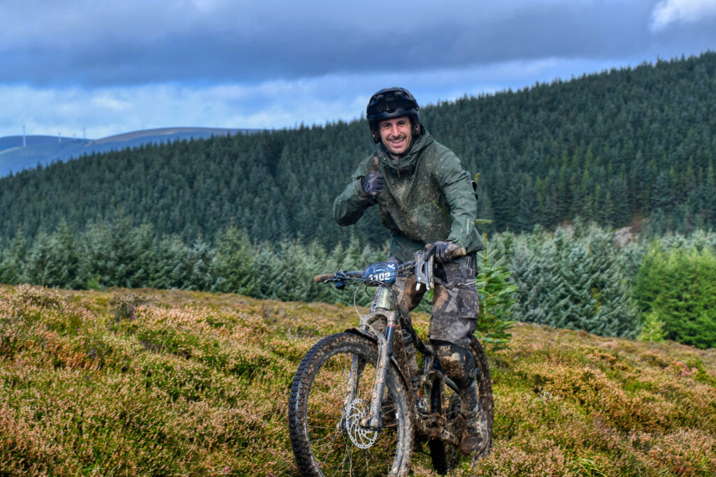 Mountain biker covered in mud, smiling and giving a thumbs-up during an off-road ride in a scenic forested landscape. Photo by Yari.